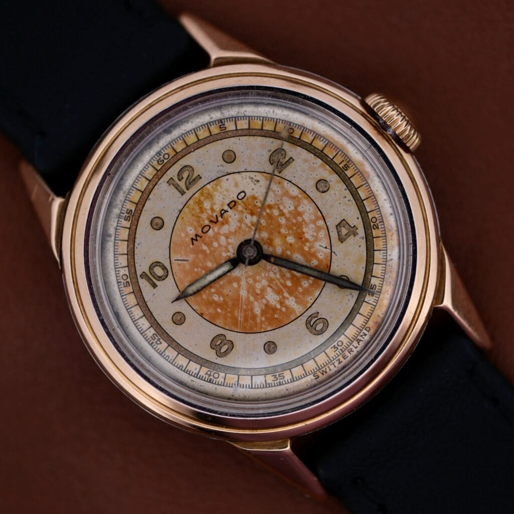 The 1945 Movado Military Dial 14k Gold Bezel Cal. 470 features a speckled, aged dial, 14k gold bezel, and is paired with a black leather strap for a timeless vintage look.