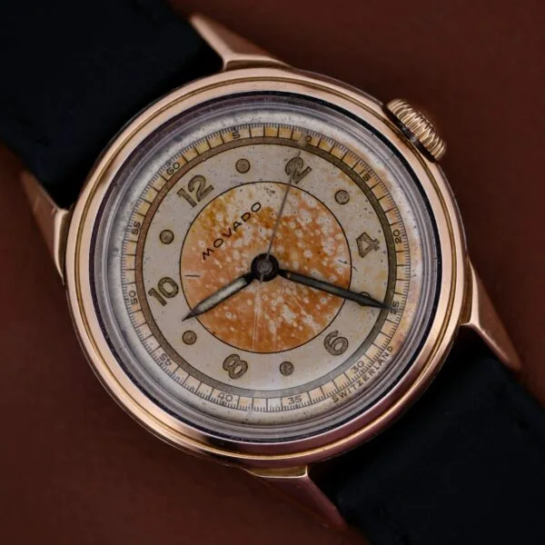 A 1945 Movado Military Dial wristwatch with a 14k gold bezel (Cal. 470), black strap, and speckled patinated dial featuring gold Arabic numerals and hands, shown on a brown background.