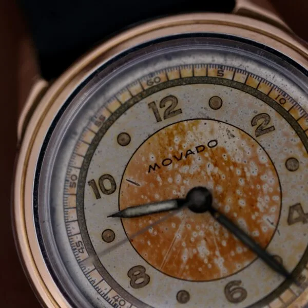 Close-up of a 1945 Movado Military Dial with a 14k gold bezel. The watch features gold-toned numerals, hands, and a black leather strap. The face shows visible signs of aging, including discoloration and spotting.