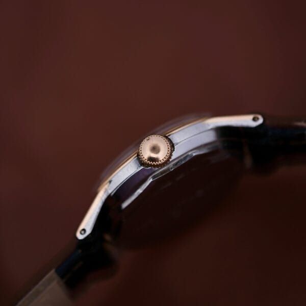 Close-up of the 1945 Movado Military Dial wristwatch displaying its centered crown against a blurred brown background. It features a dark strap and metallic casing, highlighted by a 14k gold bezel and powered by Cal. 470 movement.