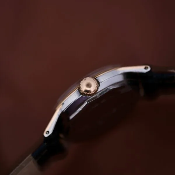 Close-up of the 1945 Movado Military Dial wristwatch displaying its centered crown against a blurred brown background. It features a dark strap and metallic casing, highlighted by a 14k gold bezel and powered by Cal. 470 movement.