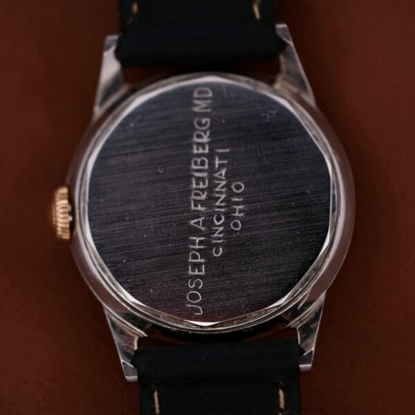 The back of a 1945 Movado Military Dial 14k Gold Bezel Cal. 470 wristwatch, featuring an engraved text: JOSEPH A. FREIBERG, MD CINCINNATI, OHIO. The watch has a black strap and is placed on a brown surface.