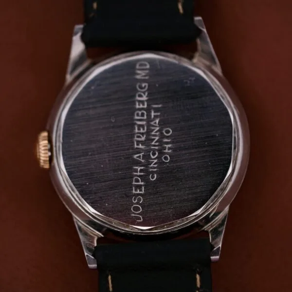 The back of a 1945 Movado Military Dial 14k Gold Bezel Cal. 470 wristwatch, featuring an engraved text: JOSEPH A. FREIBERG, MD CINCINNATI, OHIO. The watch has a black strap and is placed on a brown surface.