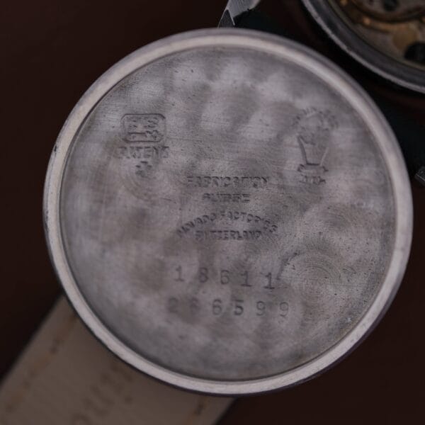 The watch case back image reveals engravings with logos, the text Fab. Suisse, Movado Factories Switzerland, serial numbers 18611 and 2665999, all on a circular brushed surface. This is from the 1945 Movado Military Dial with 14k Gold Bezel, Cal. 470.