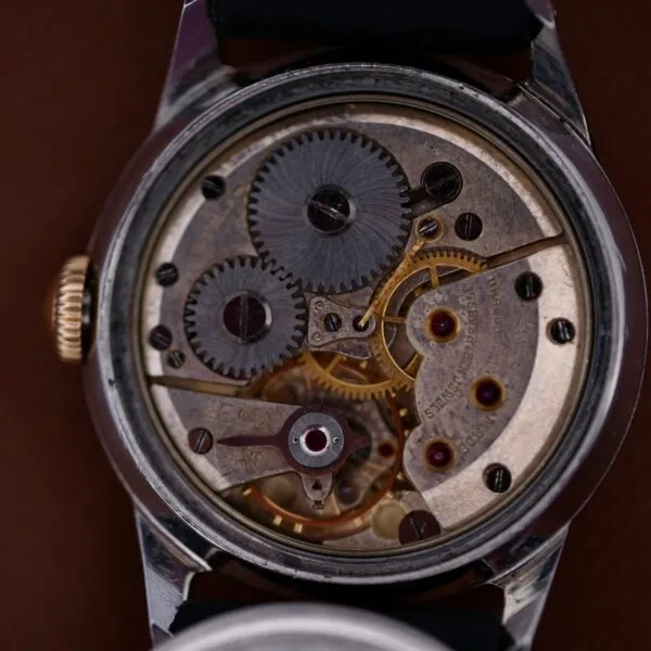 Close-up of a 1945 Movado Military Dial 14k Gold Bezel Cal. 470 watch movement with intricate gears and mechanisms, partially open metallic casing revealing various gears, visible screws, and engraved text on a brown background.