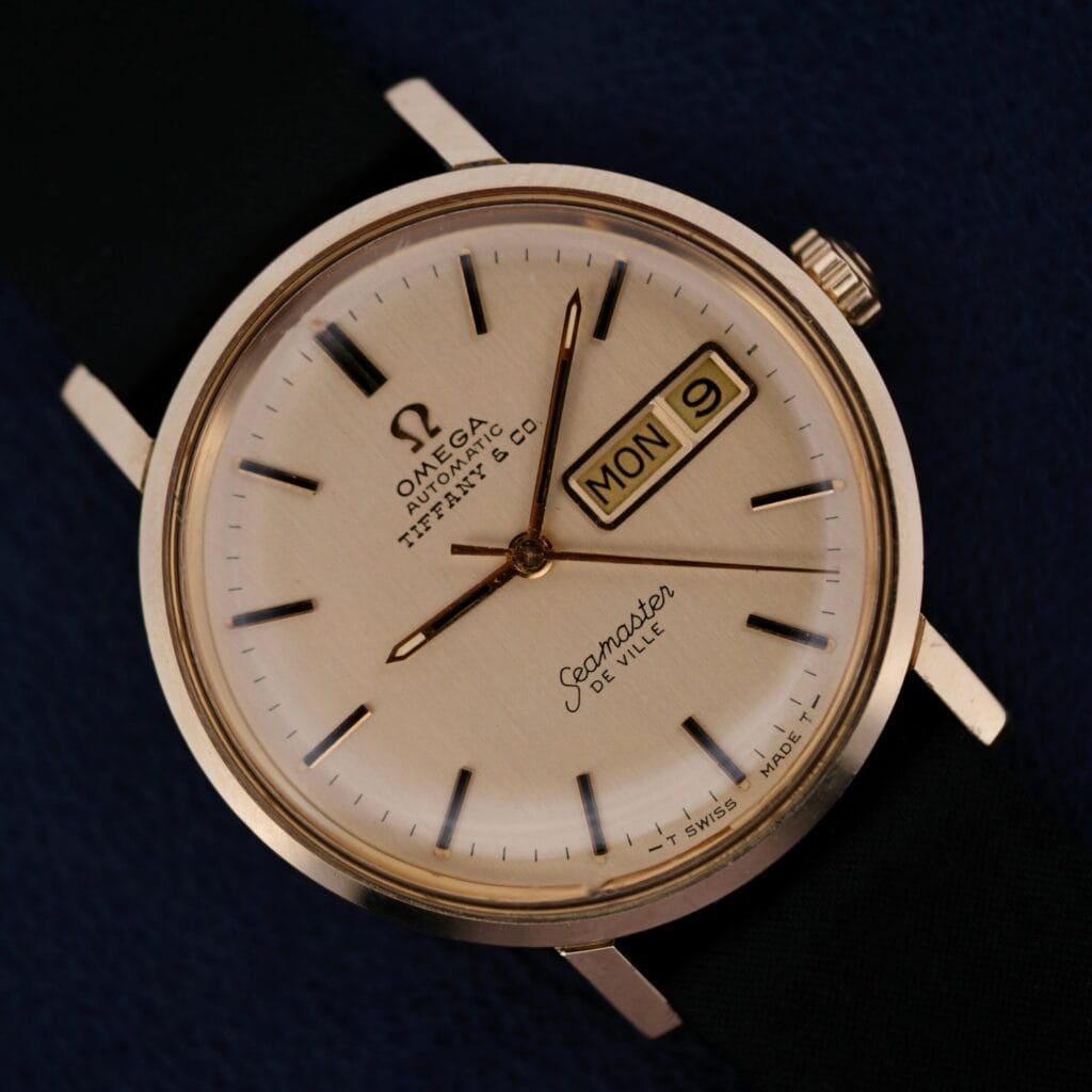 Close-up of a 1970s Omega Seamaster Deville 14k Gold Tiffany & Co. Dial C6865 watch showing Monday the 9th, with a black strap, resting on a navy surface.
