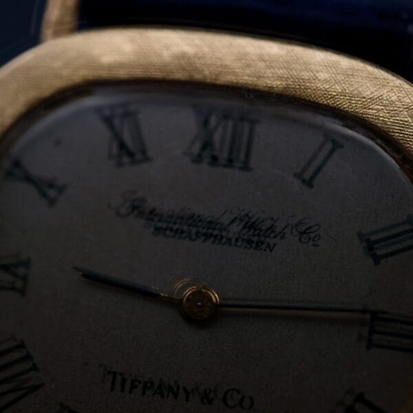 Close-up of a wristwatch face with Roman numerals, black hands, labeled Tiffany & Co. and International Watch Co. Schaffhausen, featuring a textured gold bezel. This is the IWC International Watch Co. Mens 18k Gold Tiffany & Co. Stamp Watch.