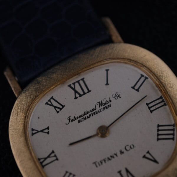 Close-up of a rectangular IWC International Watch Co. mens 18k gold Tiffany & Co. Stamp watch, featuring a white dial with black Roman numerals, slender black hands, and branding text International Watch Co Schaffhausen and Tiffany & Co, paired with a dark blue leather strap.
