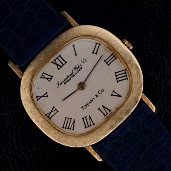 A square-faced watch with 18k gold bezel, a white dial with black Roman numerals, and the face stamped International Watch Co. Schaffhausen and Tiffany & Co. It features a dark blue textured strap.