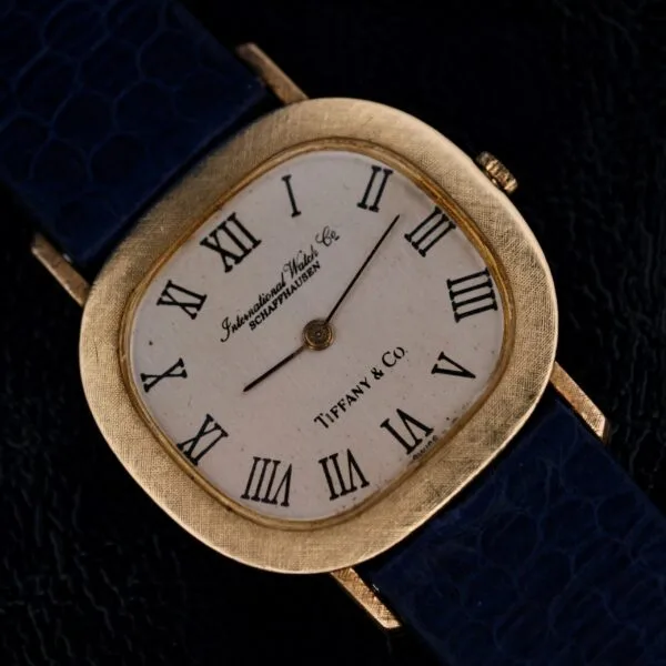 A square-faced watch with 18k gold bezel, a white dial with black Roman numerals, and the face stamped International Watch Co. Schaffhausen and Tiffany & Co. It features a dark blue textured strap.