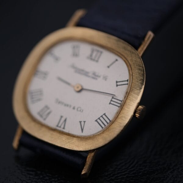 A close-up of the IWC International Watch Co. Mens 18k Gold Tiffany & Co. Stamp Watch showcases its gold rectangular case and black leather strap with a white dial featuring black Roman numerals and two central hands, positioned at a slight angle on a dark reflective surface.