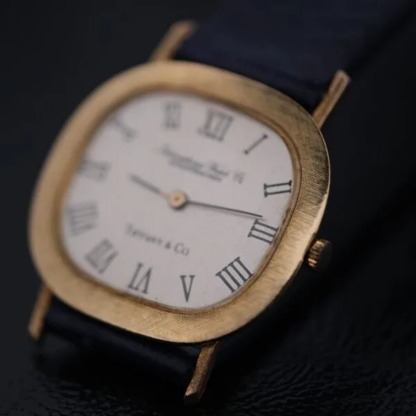 A close-up of the IWC International Watch Co. Mens 18k Gold Tiffany & Co. Stamp Watch showcases its gold rectangular case and black leather strap with a white dial featuring black Roman numerals and two central hands, positioned at a slight angle on a dark reflective surface.