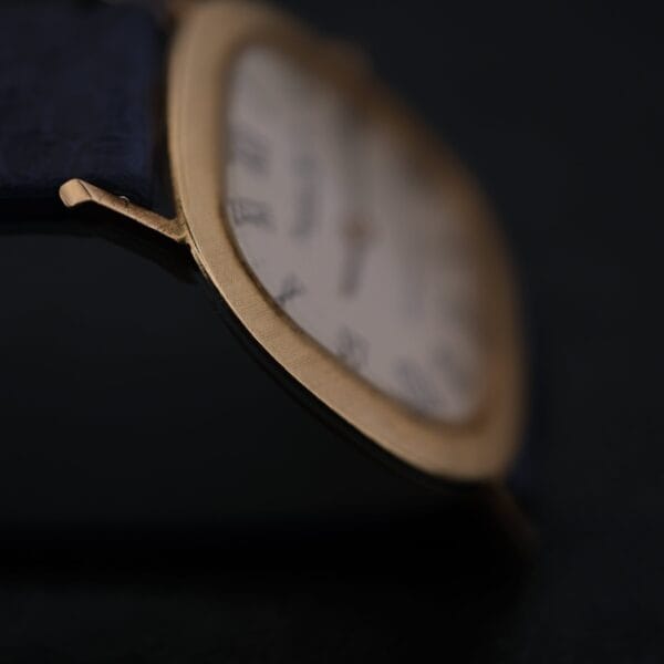 Close-up of an IWC International Watch Co. Mens 18k Gold Tiffany & Co. Stamp Watch with a rectangular gold case and Roman numeral markers on a white face, highlighting the edge and strap connection, with the face slightly blurred against a dark background.