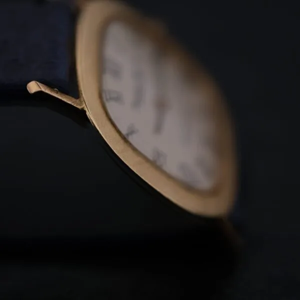 Close-up of an IWC International Watch Co. Mens 18k Gold Tiffany & Co. Stamp Watch with a rectangular gold case and Roman numeral markers on a white face, highlighting the edge and strap connection, with the face slightly blurred against a dark background.