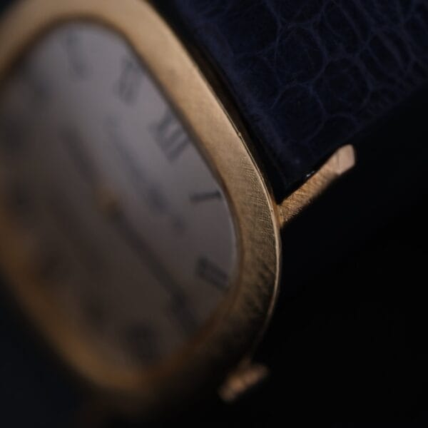 Close-up of the IWC International Watch Co. Mens 18k Gold Tiffany & Co. Stamp Watch with a textured black leather strap; its classic face displays Roman numerals, highlighted by elegant lighting that emphasizes the gold and leather textures.