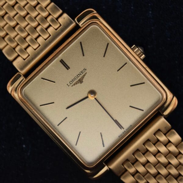 The Longines Square Case Sapphire Crown features a gold square case, gold mesh band, minimal black hour markers, and two hands on a plain gold face, showcased against a black background.