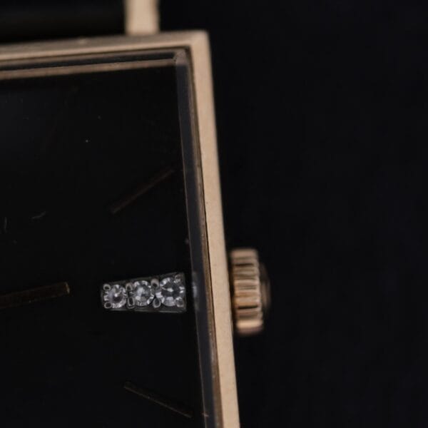 Close-up of the Omega 14K Square Case Diamond Markers showing its black watch face with minimal markings, gold-colored border, textured crown, and a silver or diamond-like hour marker.