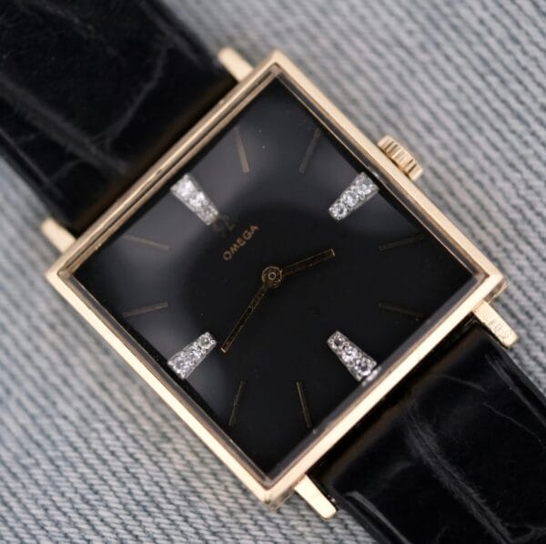 The Omega 14K Square Case Diamond Markers watch features a square gold case, black dial, black leather strap, gold hour markers and hands, with diamond accents at the 3, 6, 9, and 12 o’clock positions.