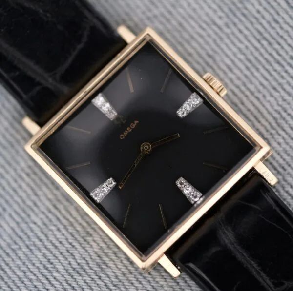 The Omega 14K Square Case Diamond Markers watch features a square gold case, black dial, black leather strap, gold hour markers and hands, with diamond accents at the 3, 6, 9, and 12 o’clock positions.