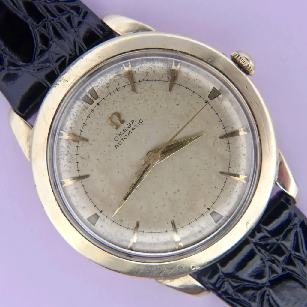 An Omega Automatic Bumper Cal. 354 wristwatch with a gold case, aged off-white dial, gold hour markers and hands, black textured leather strap, and visible wear and patina.