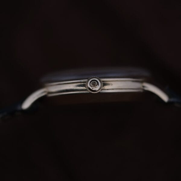 Close-up side view of the Omega Automatic Bumper Small Seconds wristwatch, featuring a metallic case and a visible crown with a circular logo. The watch face is hidden, set against a dark, blurred background.