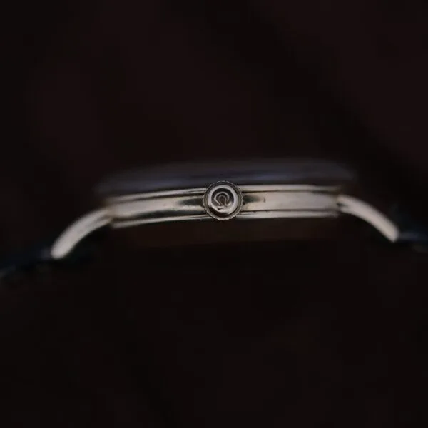 Close-up side view of the Omega Automatic Bumper Small Seconds wristwatch, featuring a metallic case and a visible crown with a circular logo. The watch face is hidden, set against a dark, blurred background.