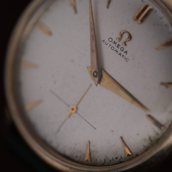 A close-up of the Omega Automatic Bumper Small Seconds wristwatch face, featuring gold hour markers and hands with visible dust and wear. The time shown is around 4:07.