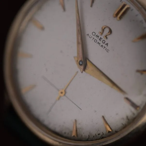 A close-up of the Omega Automatic Bumper Small Seconds wristwatch face, featuring gold hour markers and hands with visible dust and wear. The time shown is around 4:07.