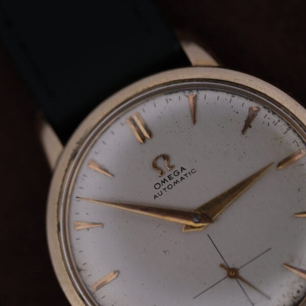 A close-up view of the Omega Automatic Bumper Small Seconds wristwatch featuring a white face, gold hour markers and hands, black strap, and visible Omega logo with Automatic on the dial.