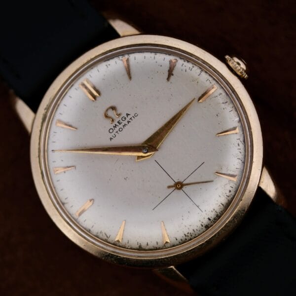 A close-up of an Omega Automatic Bumper Small Seconds wristwatch with a gold case, white dial, gold hour markers, black leather strap, and visible aging on the face. The time displayed is about 8:21.