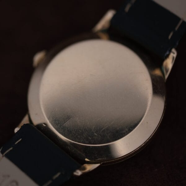 A close-up of the back of an Omega Automatic Bumper Small Seconds wristwatch with a metal case and dark straps, showing faint scratches on the surface and no visible markings or engravings.