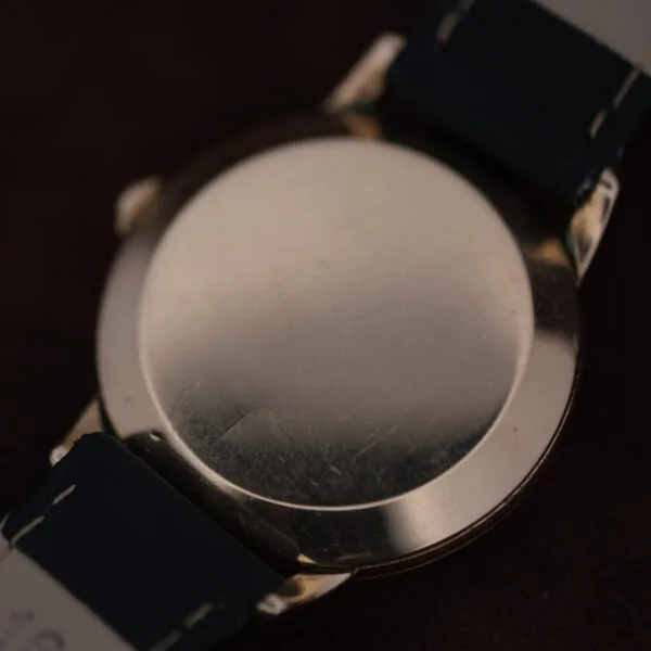 A close-up of the back of an Omega Automatic Bumper Small Seconds wristwatch with a metal case and dark straps, showing faint scratches on the surface and no visible markings or engravings.
