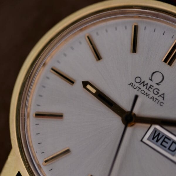 Close-up of a gold Omega Automatic Day/Date Sunburst Cal. 1020 watch, displaying the white face with black and gold hour markers, visible Omega logo, and partly shown day-date display on the right.