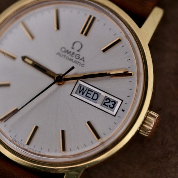 Close-up of an Omega Automatic Day/Date Sunburst Cal. 1020 watch showing Wednesday, the 23rd. It features a white face with black and gold hour markers and hands, and a partially visible brown leather strap.