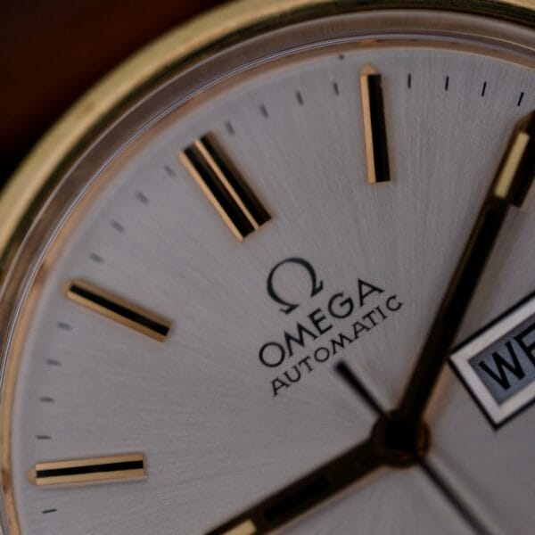 Close-up of an Omega Automatic Day/Date Sunburst Cal. 1020 watch face with a silver dial, gold hour markers and hands, a visible logo, the word Automatic, and a date window displaying WED on the right side. It has a classic and elegant design.