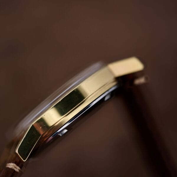 Close-up of the Omega Automatic Day/Date Sunburst Cal. 1020 watch, featuring a gold-colored metal case and glass face on a brown leather strap, with a blurred brown background.