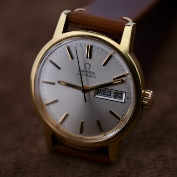 A gold-colored Omega Automatic Day/Date Sunburst Cal. 1020 wristwatch with a brown leather strap features a silver face, black hour markers, two hands, and the brand logo. The date window displays WED 23, and it rests on a soft brown surface.
