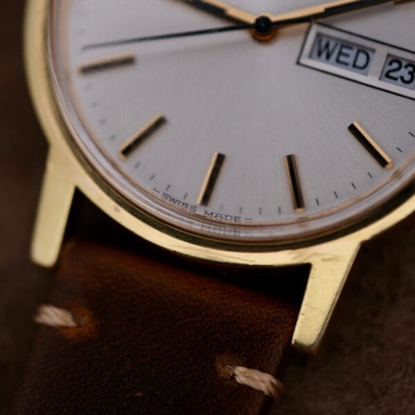 Close-up of an Omega Automatic Day/Date Sunburst Cal. 1020 with a gold-toned case and leather strap. It features minimalist hour markers, black hands, and a WED 23 day/date window. The SWISS MADE mark is at the bottom, against a textured fabric background.