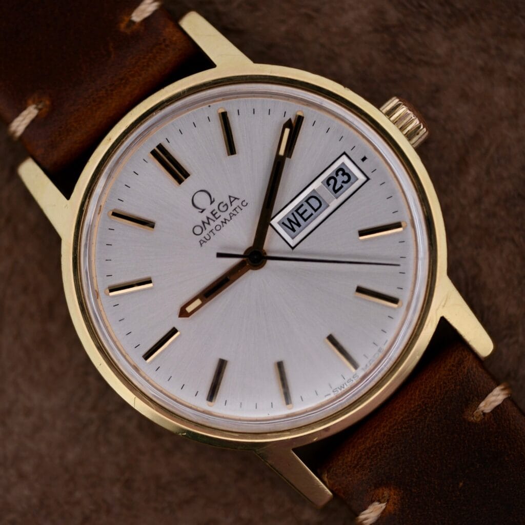 The Omega Automatic Day/Date Sunburst Cal. 1020 features a gold case, brown leather strap, automatic movement, and classic Day Date display (WED 23).