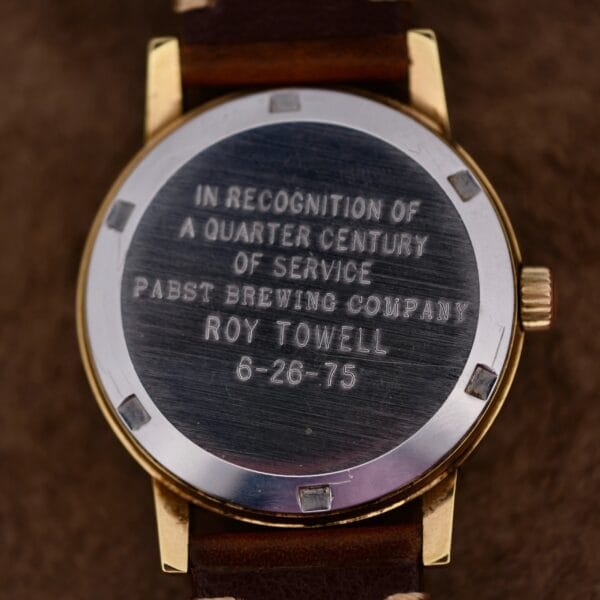 The image displays the back of an Omega Automatic Day/Date Sunburst Cal. 1020 gold watch with a brown leather strap, engraved with: In recognition of a quarter century of service Pabst Brewing Company Roy Towell 6-26-75.