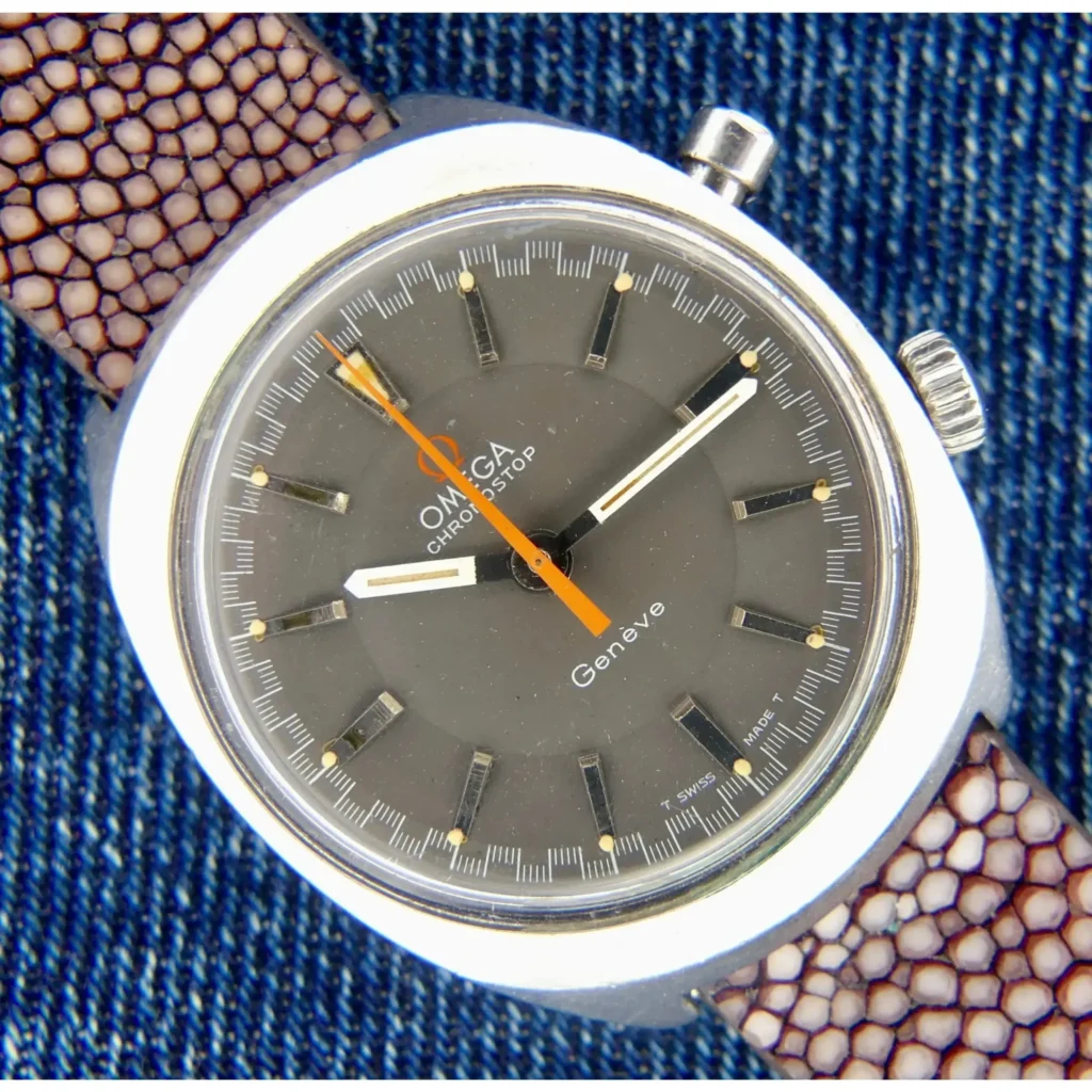 A close-up of the Omega Chronostop Ref 145.009 Caliber 865 wristwatch featuring a brown patterned strap, gray dial, silver case, orange second hand, and white hour and minute hands on blue denim fabric.