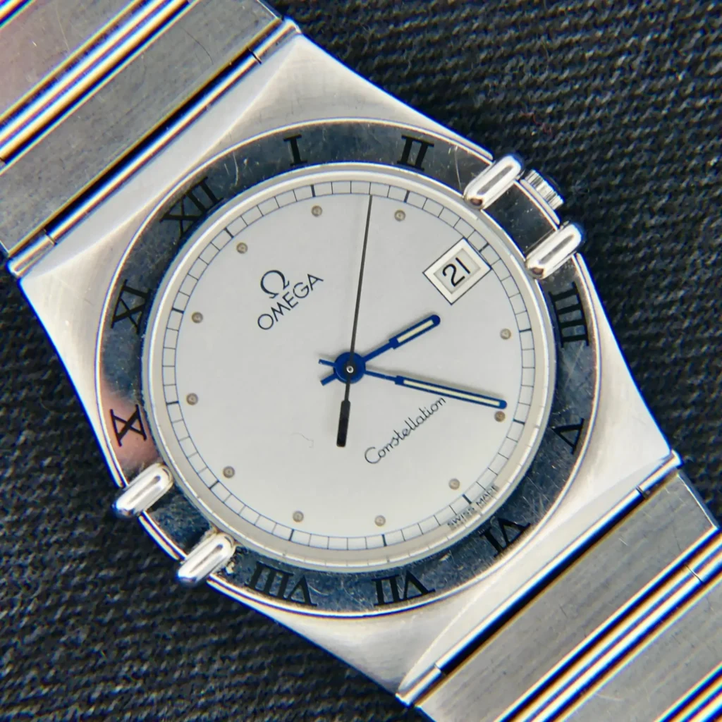 An Omega Constellation Ref. 396.10701 wristwatch with a white dial, black Roman numerals, date window at 3 o’clock, and a silver metal strap is displayed on a dark textured surface.