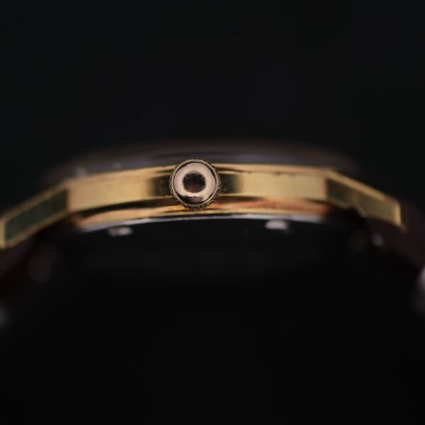 Close-up side view of an Omega Geneve Automatic Date gold wristwatch crown against a dark, blurred background, highlighting the crown’s round end and the watch casing’s metallic finish.