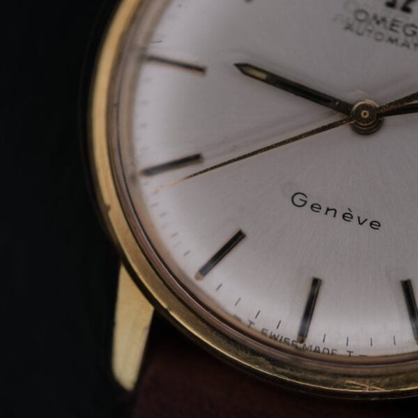 Close-up of an Omega Geneve Automatic Date wristwatch, highlighting the gold case, silver dial, black hour markers and hands, with “Genève” and “Swiss Made” visible on the face.