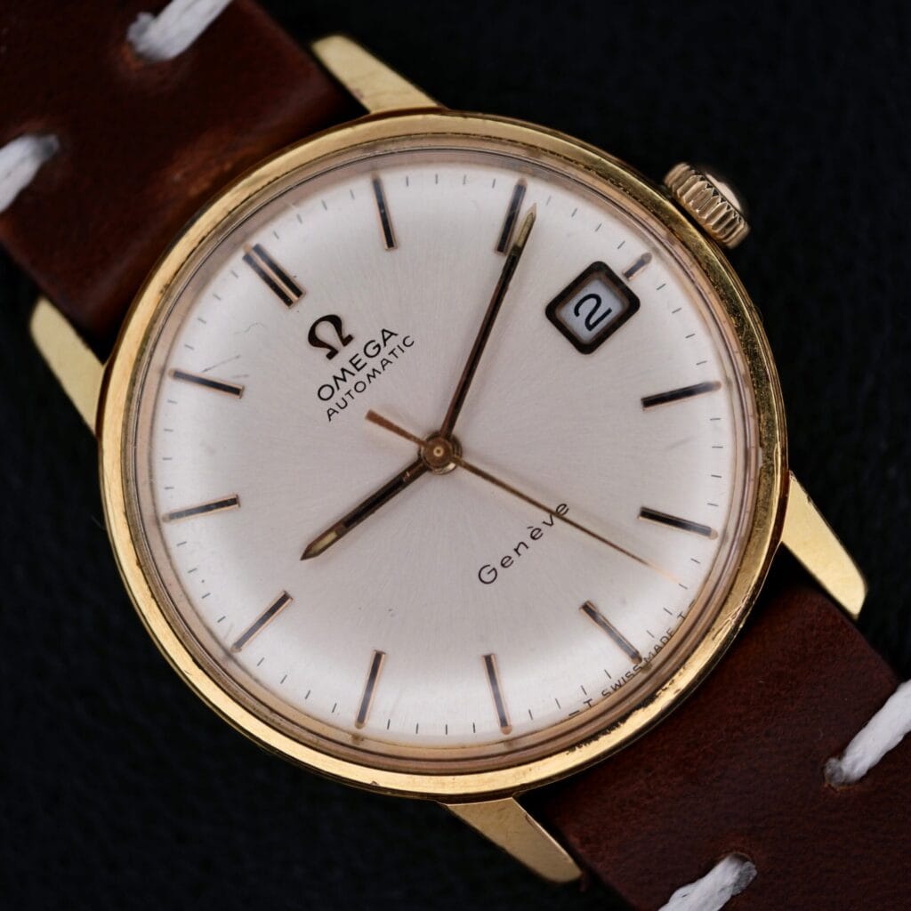 An Omega Geneve Automatic Date wristwatch with a gold case, white dial, date at 3 o’clock (showing “2”), brown leather strap with white stitching, and slim markers, is photographed against a dark background.