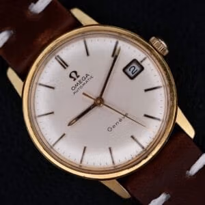 An Omega Geneve Automatic Date wristwatch with a gold case, white dial, date at 3 o’clock (showing “2”), brown leather strap with white stitching, and slim markers, is photographed against a dark background.