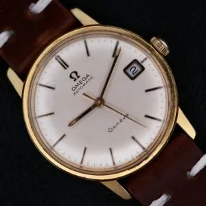 An Omega Geneve Automatic Date wristwatch with a gold case, white dial, date at 3 o’clock (showing “2”), brown leather strap with white stitching, and slim markers, is photographed against a dark background.