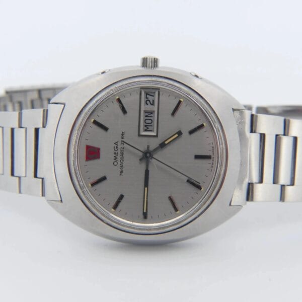 The Omega Megaquartz 32 KHz Day/Date Cal. 1310 is a silver wristwatch with a metal strap and round face, showcasing the Omega logo, Megaquartz 32 kHz, day/date windows on the right, black hands/indices, and a red accent at 12 oclock.
