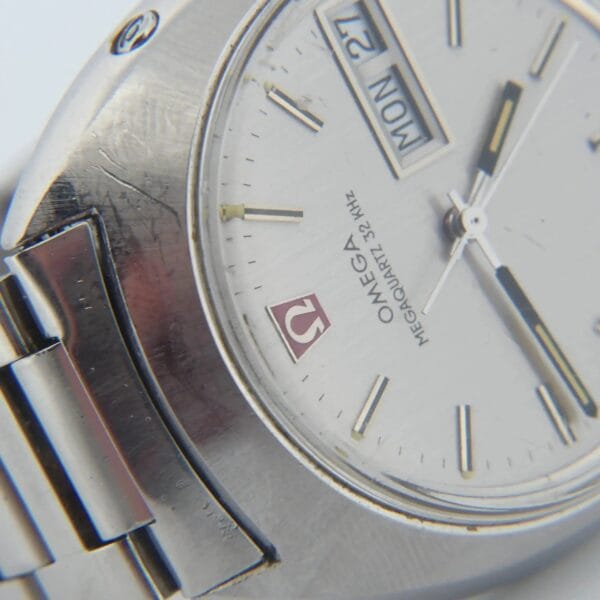 Close-up of a silver Omega Megaquartz 32 KHz Day/Date Cal. 1310 wristwatch with metal bracelet, featuring MON and 21 on the display and OMEGA on the dial; bezel shows wear.