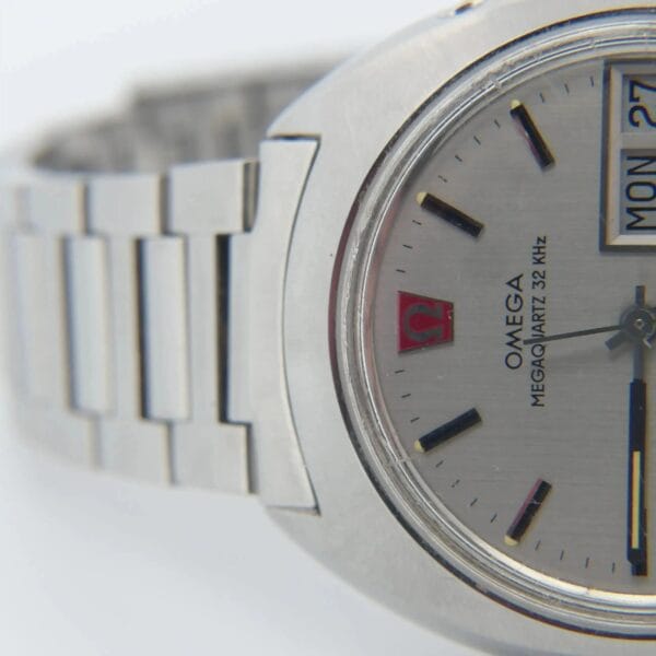 Close-up of the Omega Megaquartz 32 KHz Day/Date Cal. 1310 wristwatch with a silver metal band, featuring a round face with black hour markers, day and date display, red Omega logo, and MEGAQUARTZ 32 KHZ text on the dial.